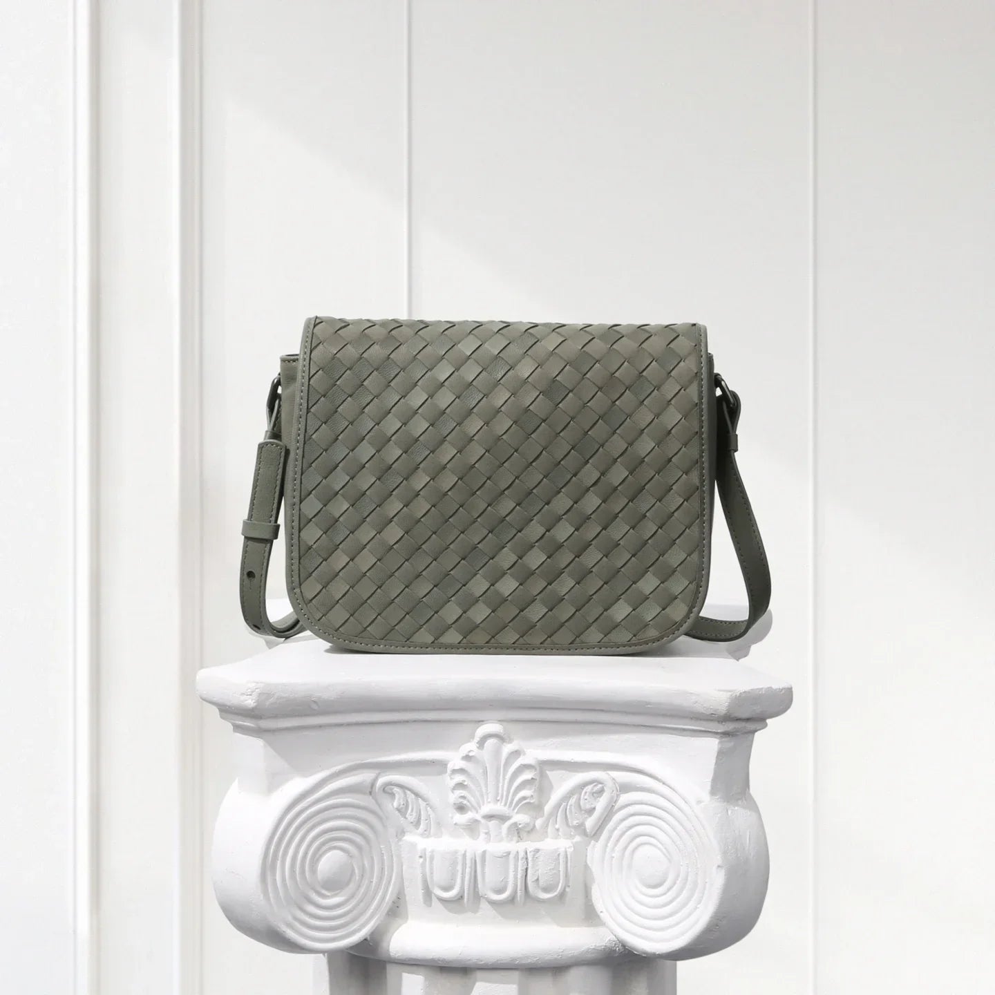 Gray woven handbag on a decorative white pedestal against a light wooden panel background