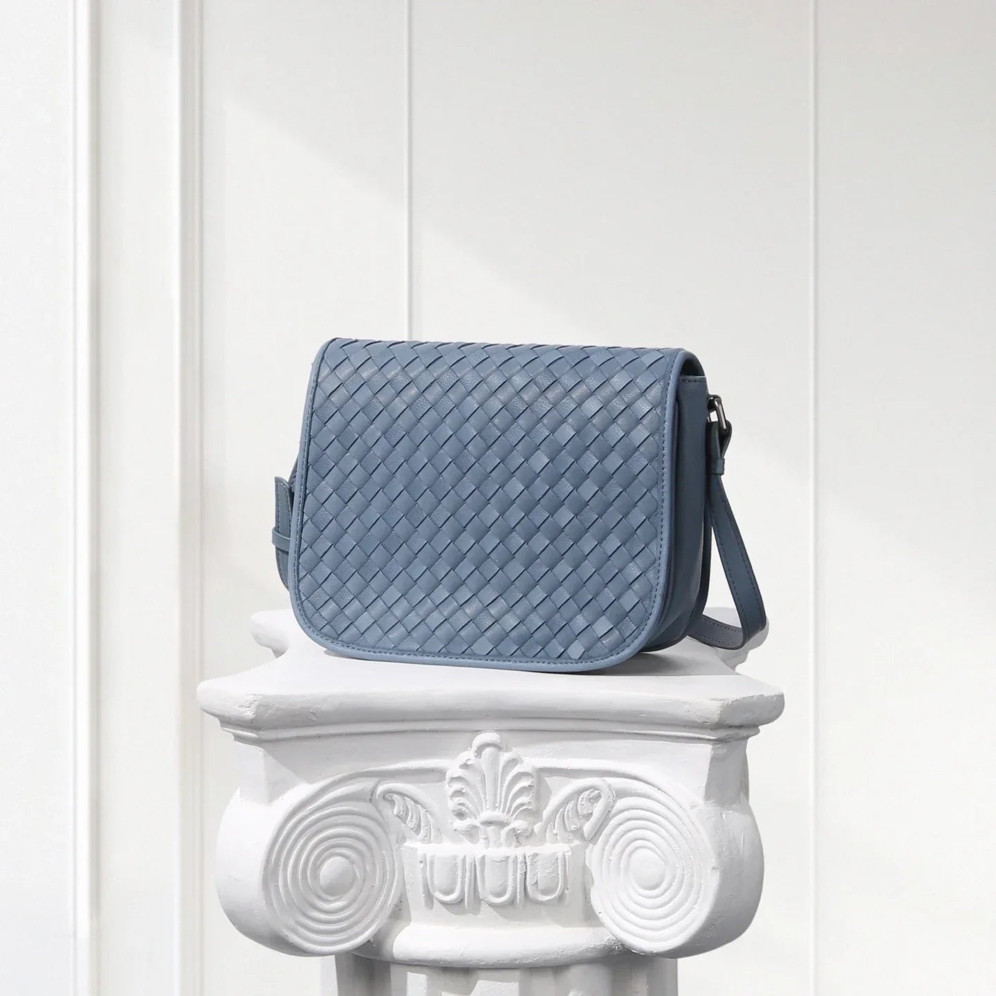 Blue woven handbag on a decorative white pedestal against a white wooden panel background
