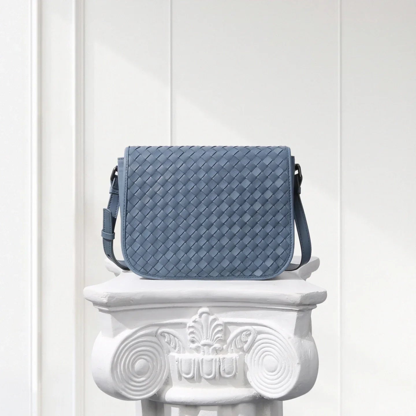 Blue woven handbag on a decorative white pedestal against a light gray background