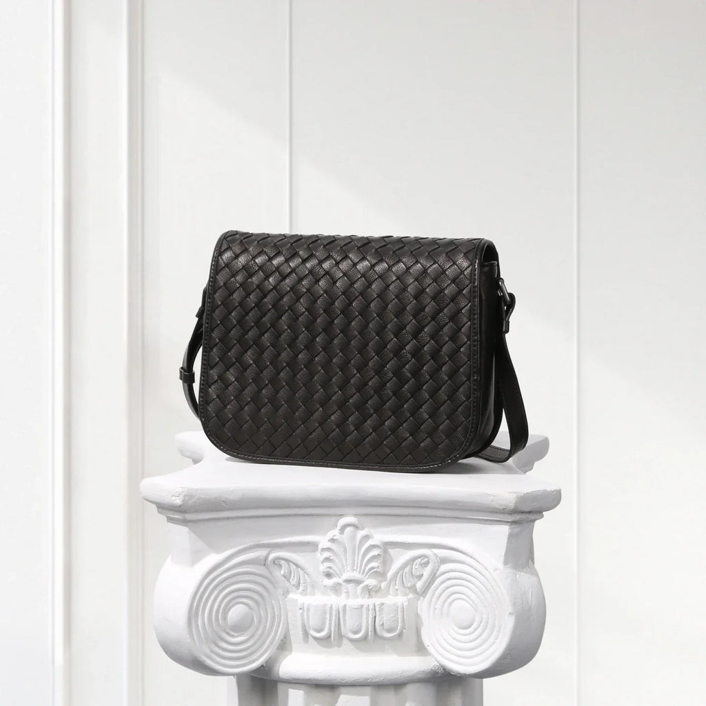 Black woven handbag on a decorative white pedestal against a light wooden panel background