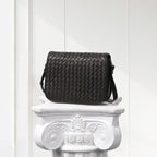 Black woven handbag on a decorative white pedestal against a light wooden panel background