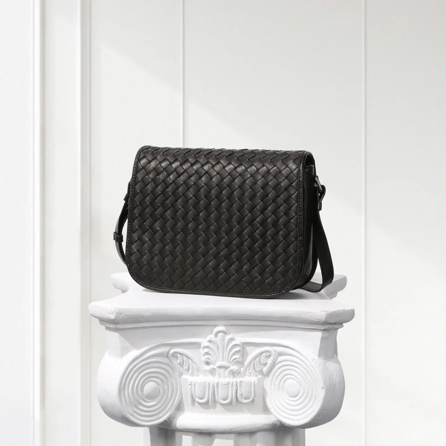 Black woven handbag on a decorative white pedestal against a light wooden panel background