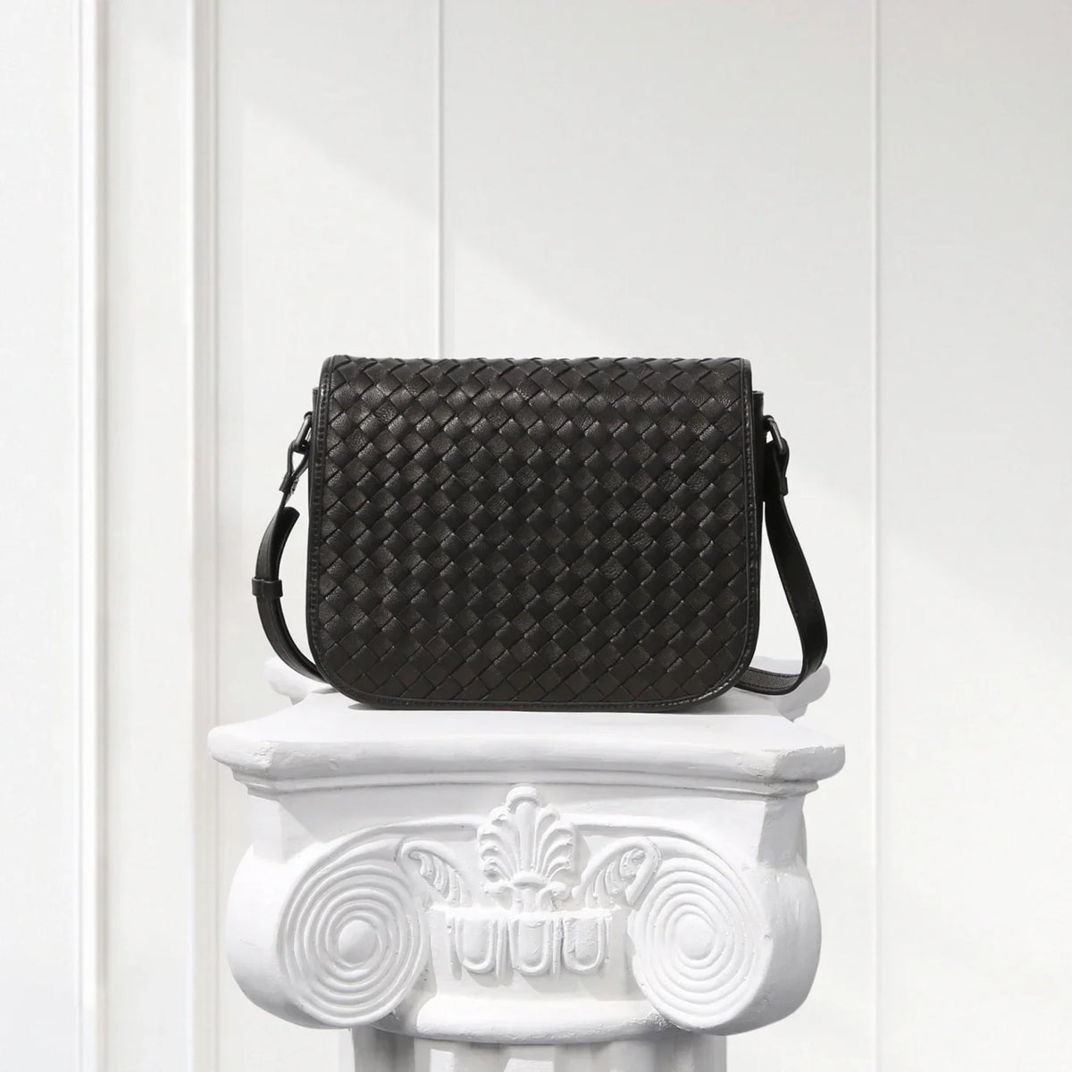 Black woven handbag on a decorative white pedestal against a light background