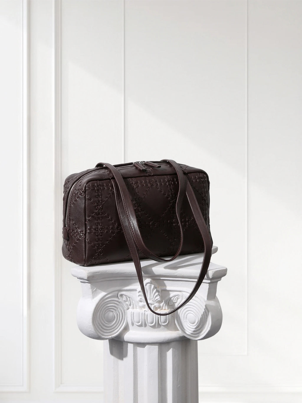 Brown textured handbag on a decorative white column against a white paneled wall