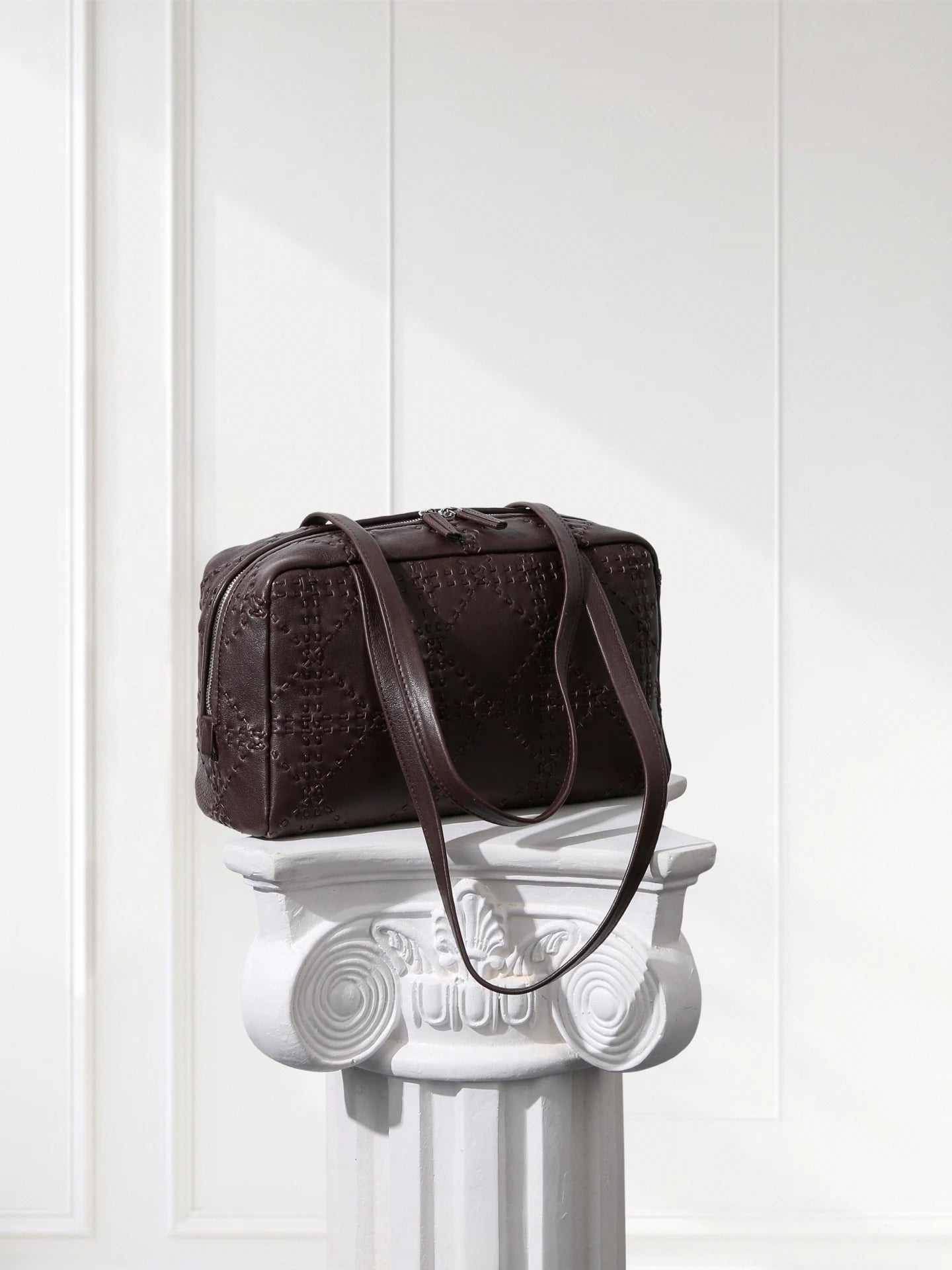 Brown textured handbag on a decorative white column against a white paneled wall