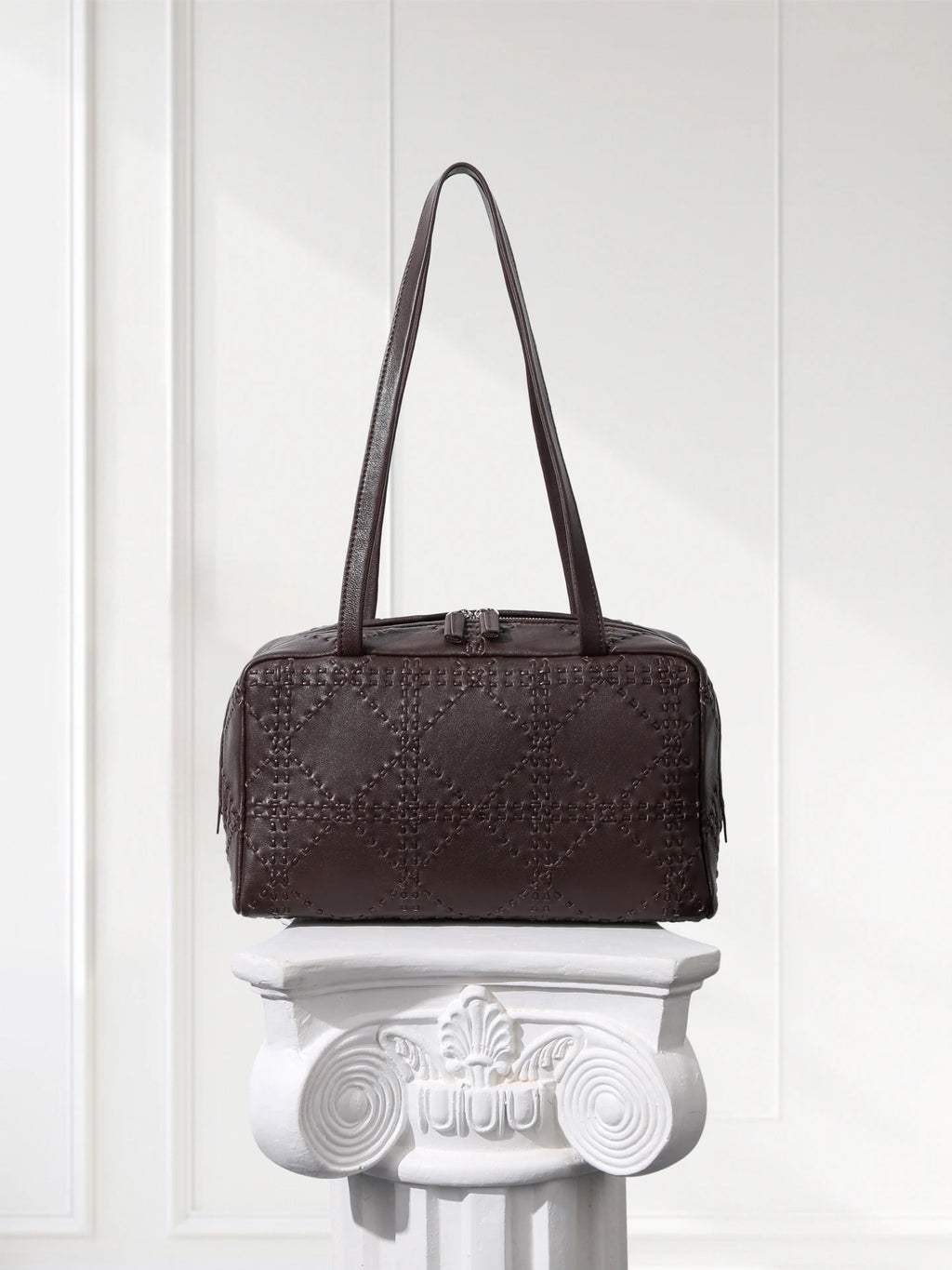 Brown textured handbag on a decorative white pedestal against a light background