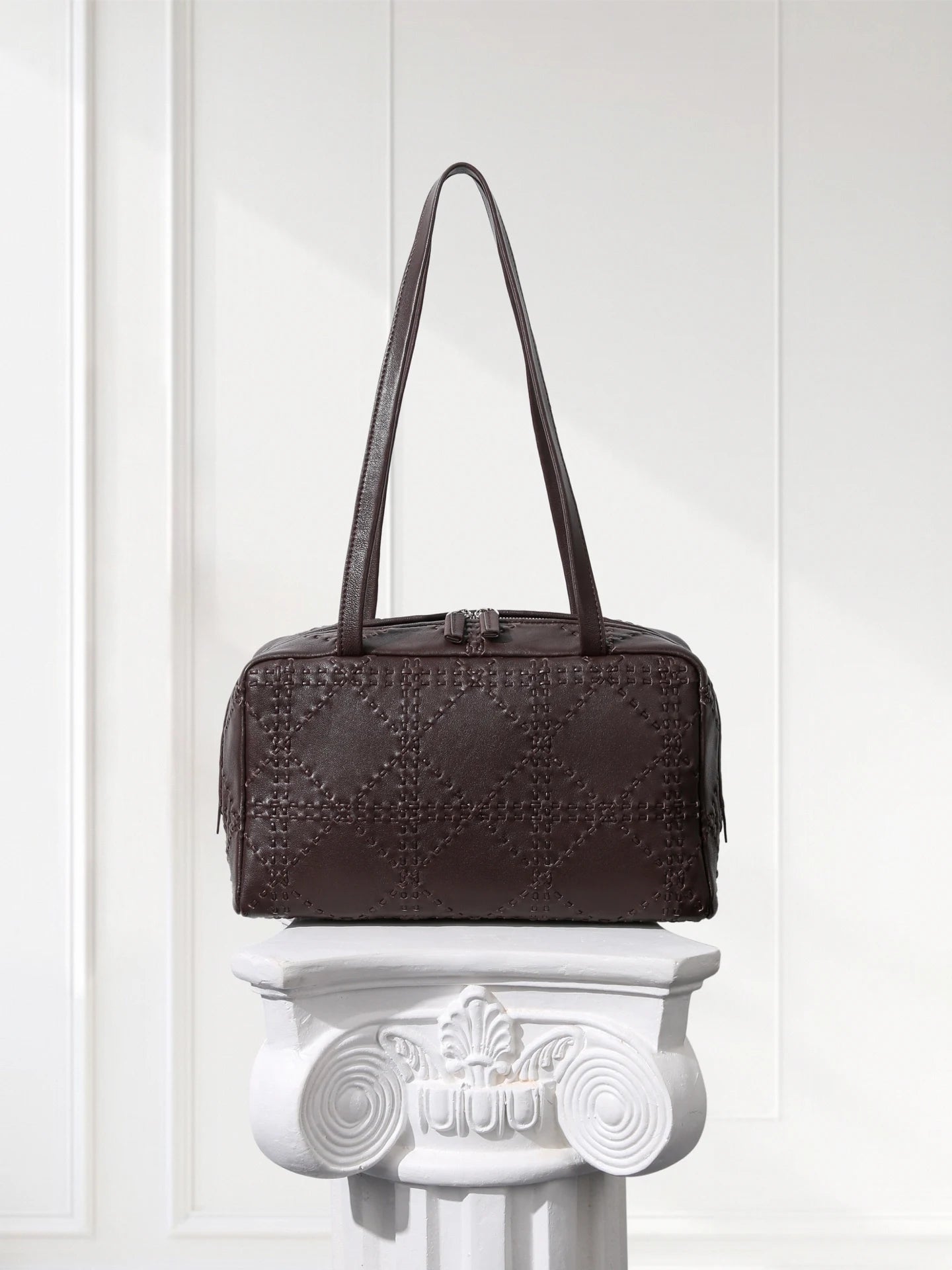 Brown textured handbag on a decorative white pedestal against a light background
