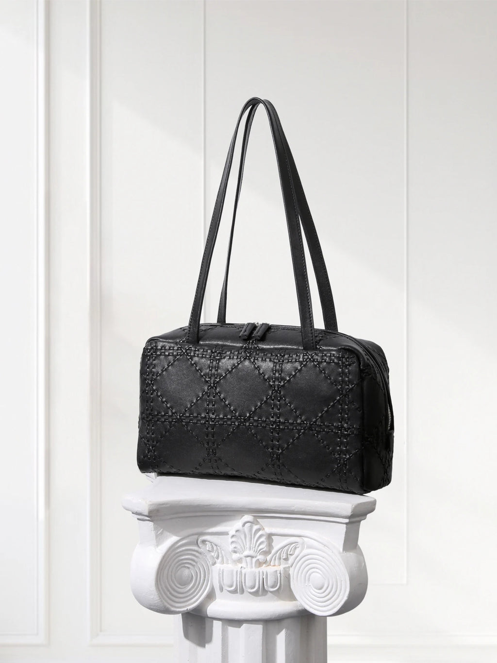Black handbag on a decorative white pedestal against a white paneled wall.