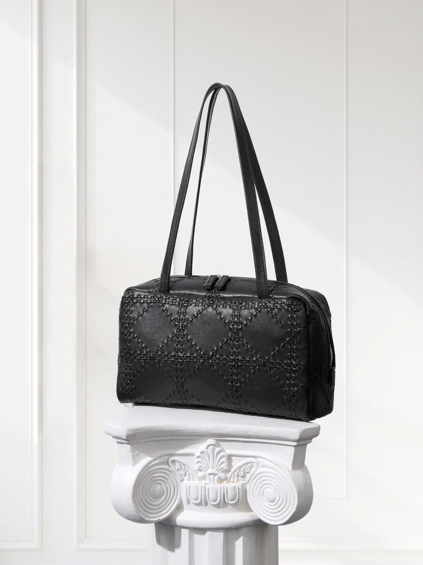 Black handbag on a decorative white pedestal against a white paneled wall.