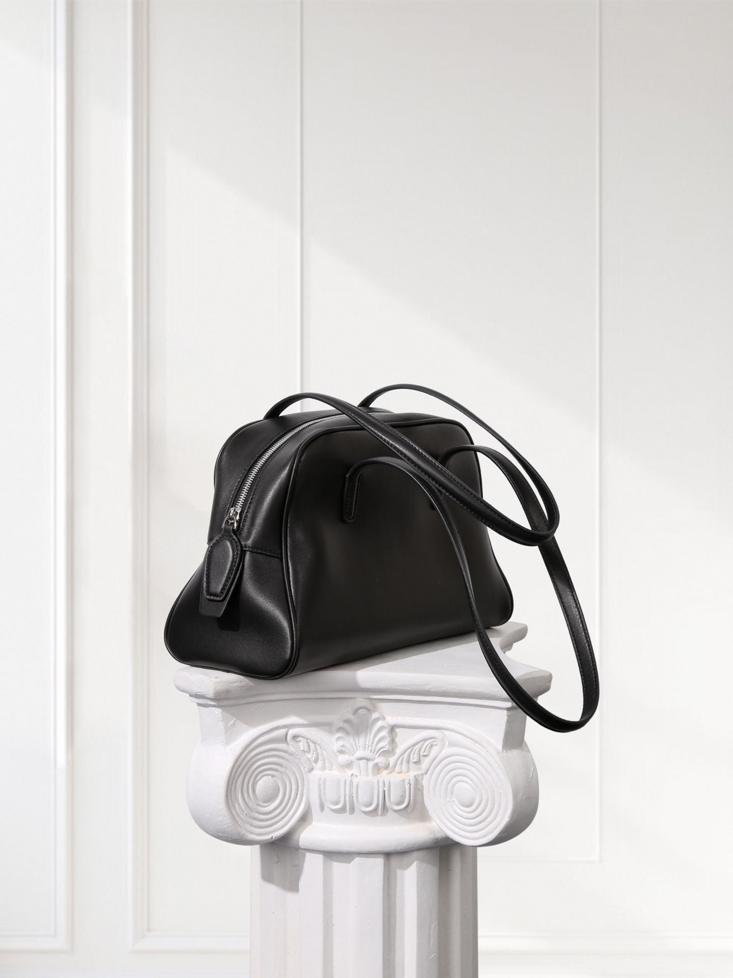Black handbag on a decorative white pedestal against a white paneled wall