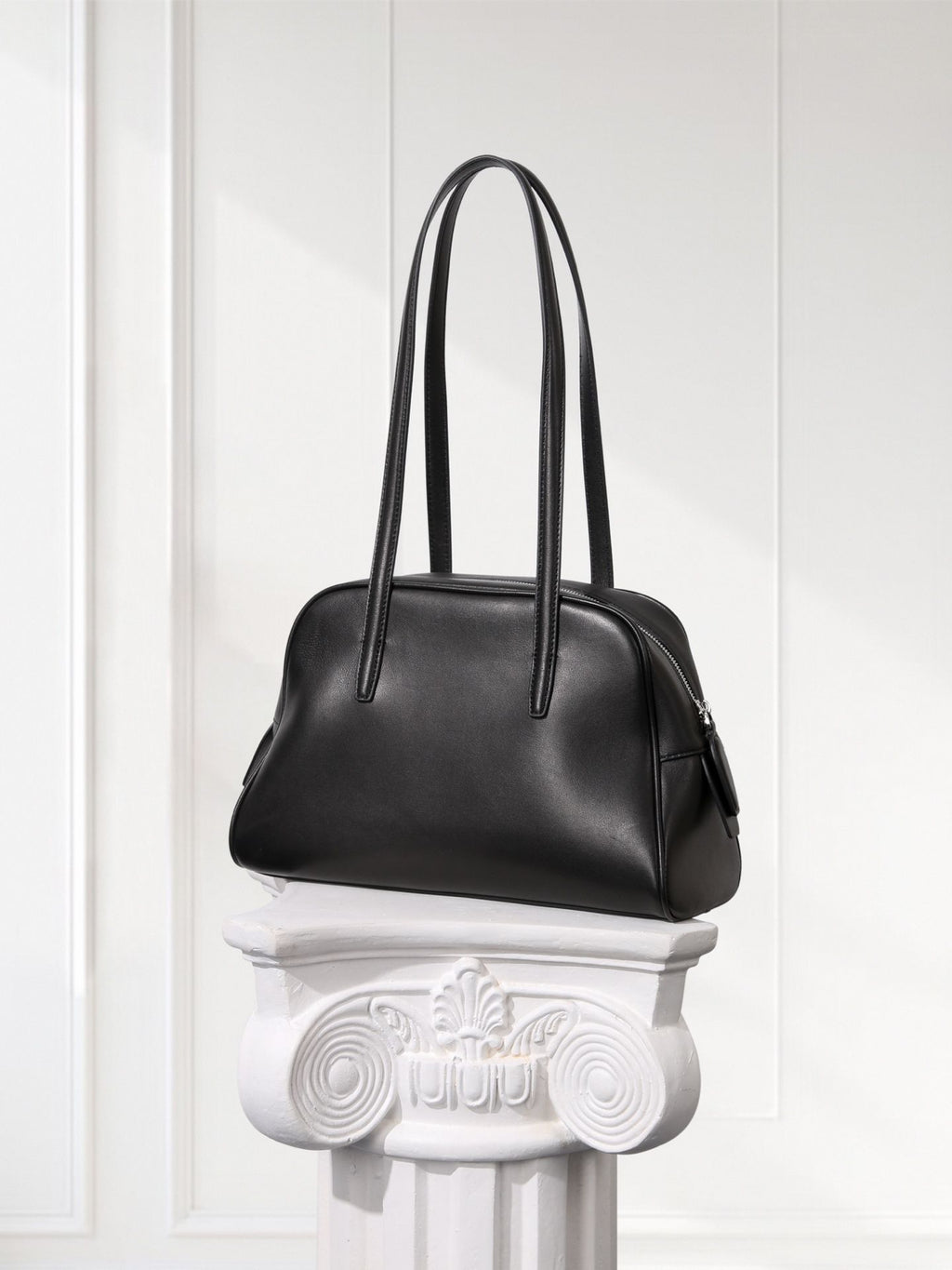 Black handbag on a decorative white pedestal against a white paneled wall.