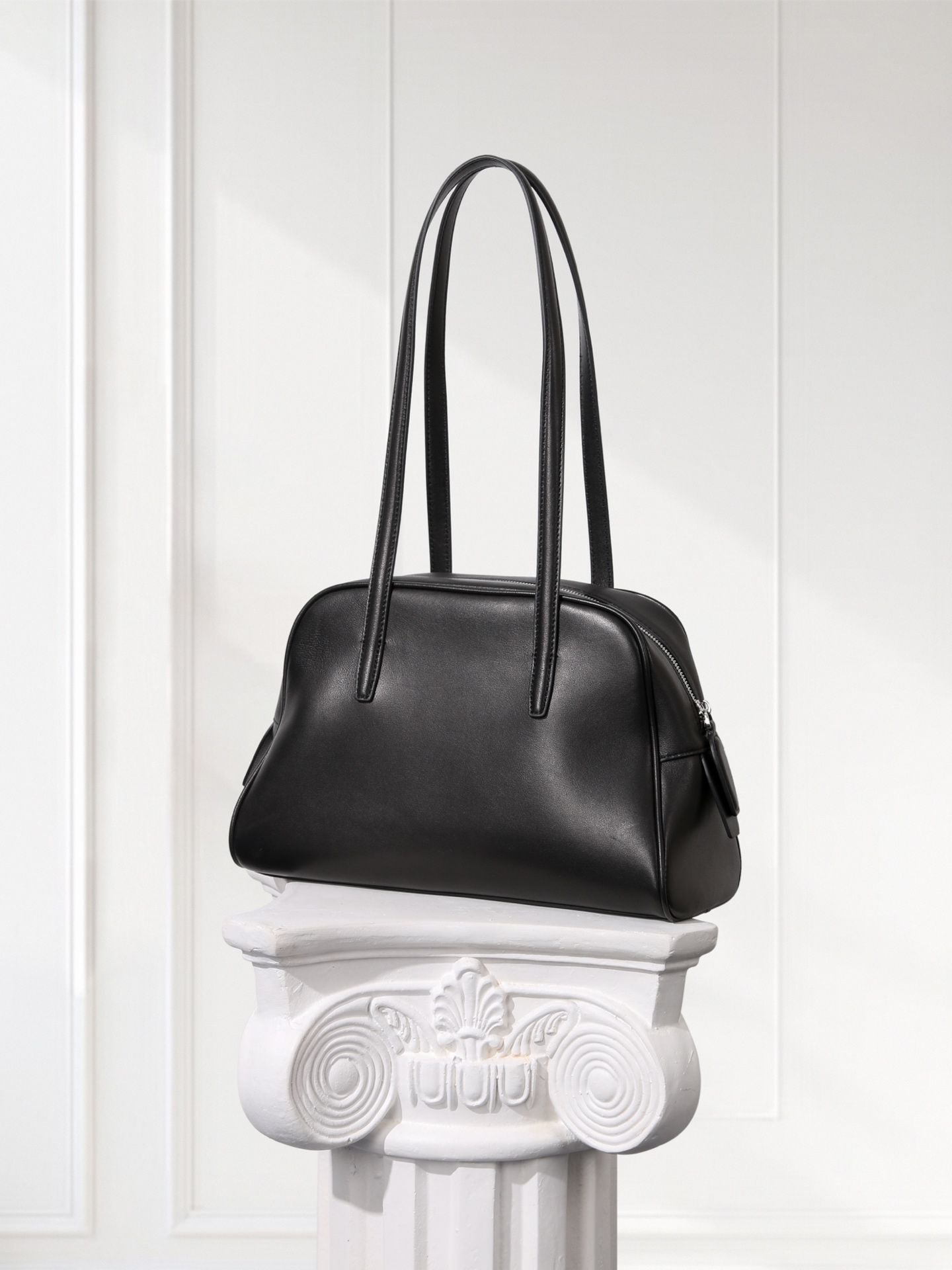 Black handbag on a decorative white pedestal against a white paneled wall.