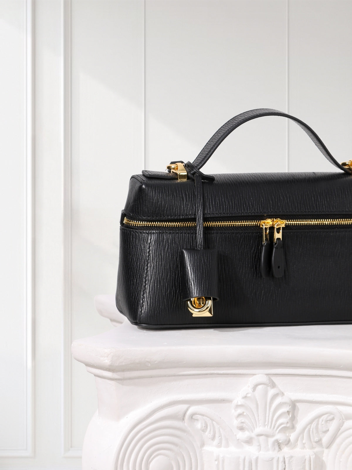 A black textured leather top-handle handbag with gold-tone hardware, resting on a white decorative pedestal against a white paneled wall.