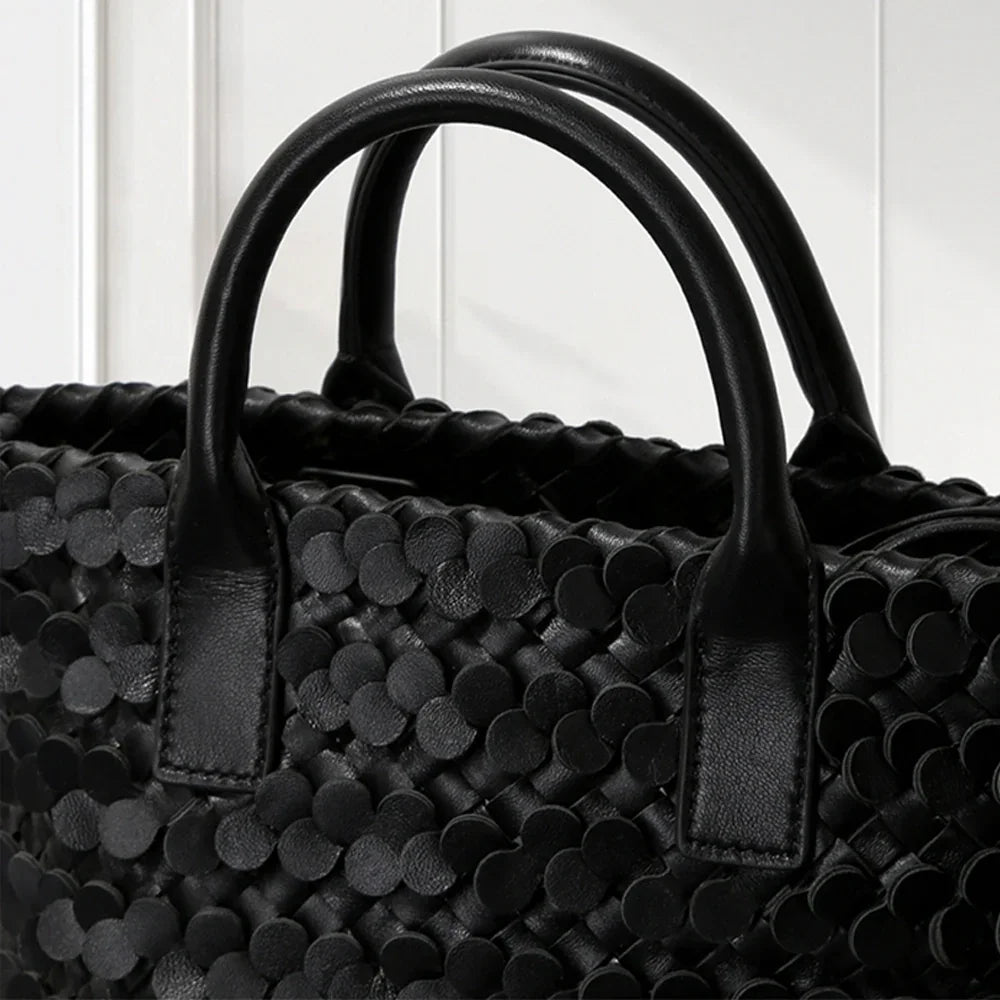 BLACK leather tote bag with circular appliqué design, placed on a decorative white pedestal.