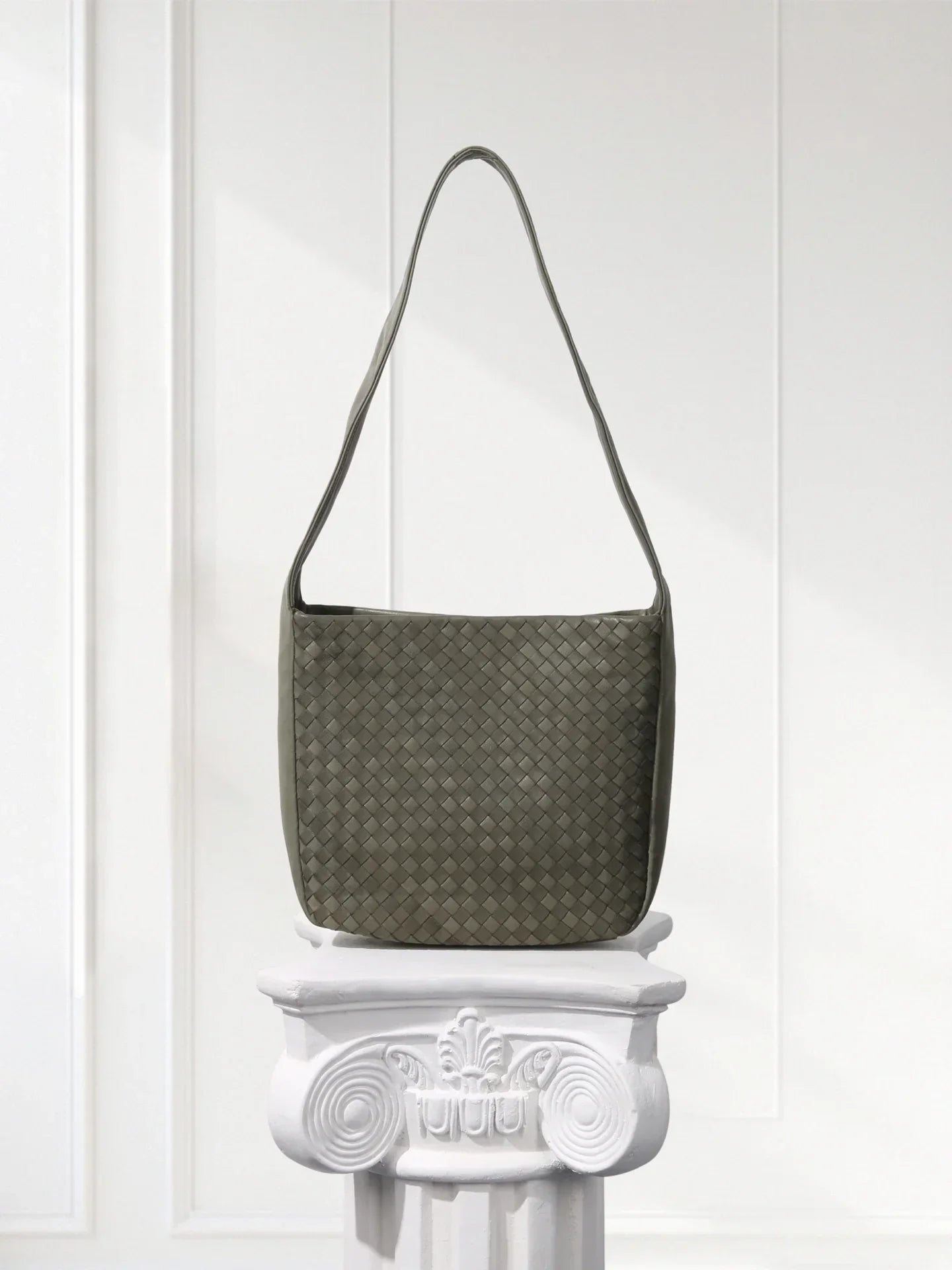 Green textured handbag on a decorative white pedestal against a white wall.