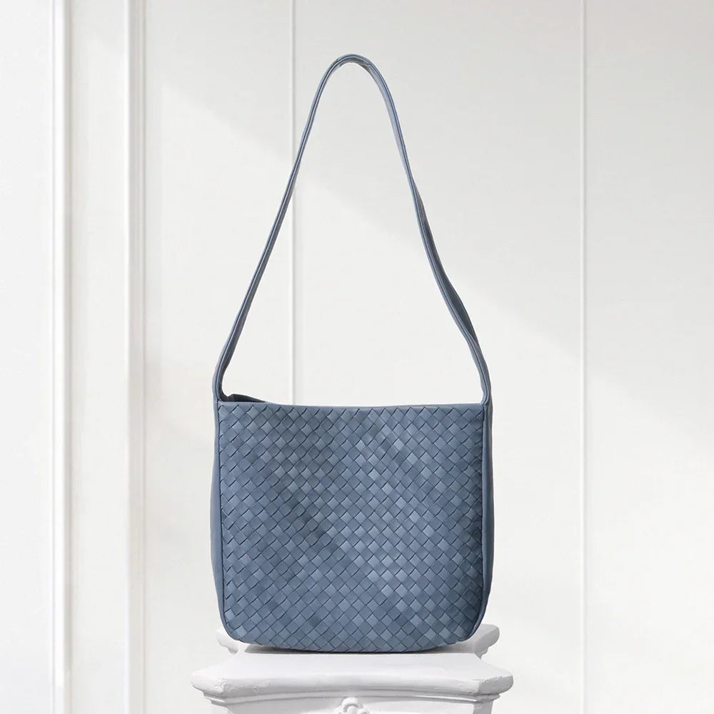 Blue woven handbag on a white pedestal against a white wooden panel background