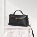 A structured black leather handbag with a top handle and gold-tone hardware, displayed on a white decorative pedestal against a white paneled wall.
