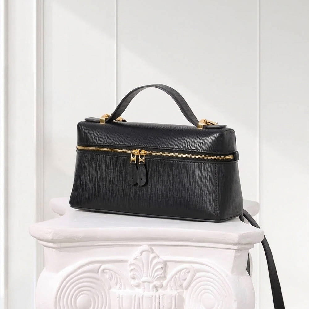 A structured black leather handbag with a top handle and gold-tone hardware, displayed on a white decorative pedestal against a white paneled wall.
