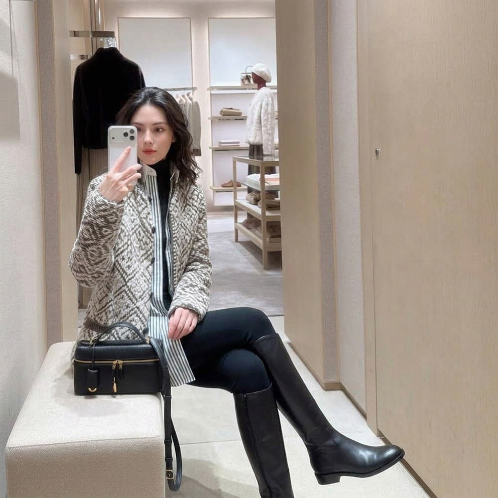 A woman takes a mirror selfie in a boutique while wearing a beige and black geometric patterned quilted jacket, a black turtleneck, a striped shirt, black leggings, and black knee-high boots. She is seated on a beige bench next to a black handbag.