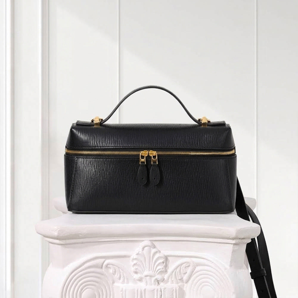 A black textured leather handbag with gold-tone hardware, a top handle, and a front zip closure, displayed on a white decorative pedestal against a white paneled wall.