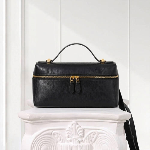 A black textured leather handbag with gold-tone hardware, a top handle, and a front zip closure, displayed on a white decorative pedestal against a white paneled wall.
