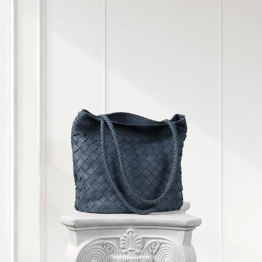 Blue woven handbag on a decorative white pedestal against a white paneled wall.