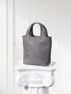 Gray handbag with woven pattern on a decorative white pedestal against a white wall.