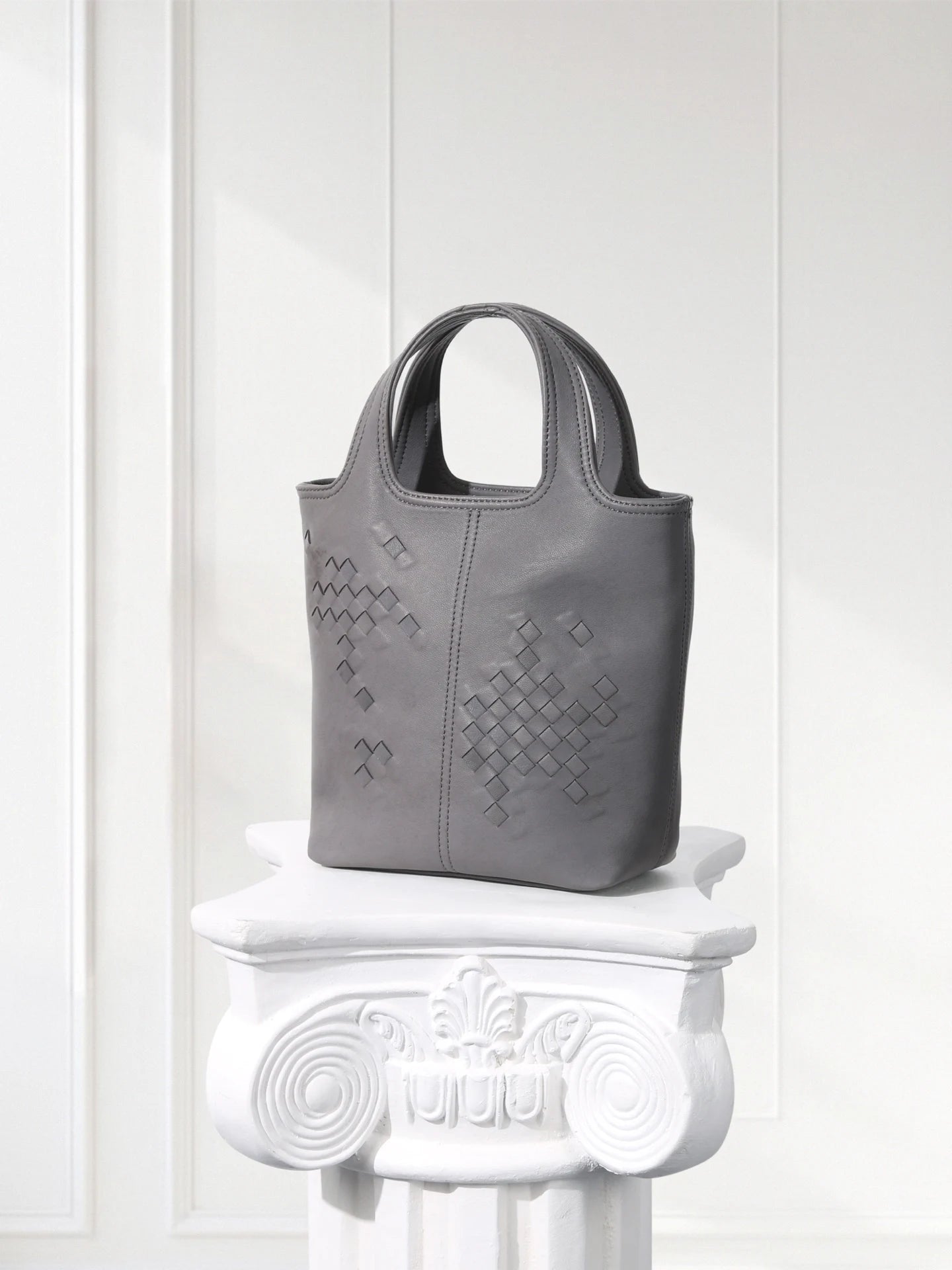 Gray handbag with woven pattern on a decorative white pedestal against a white wall.