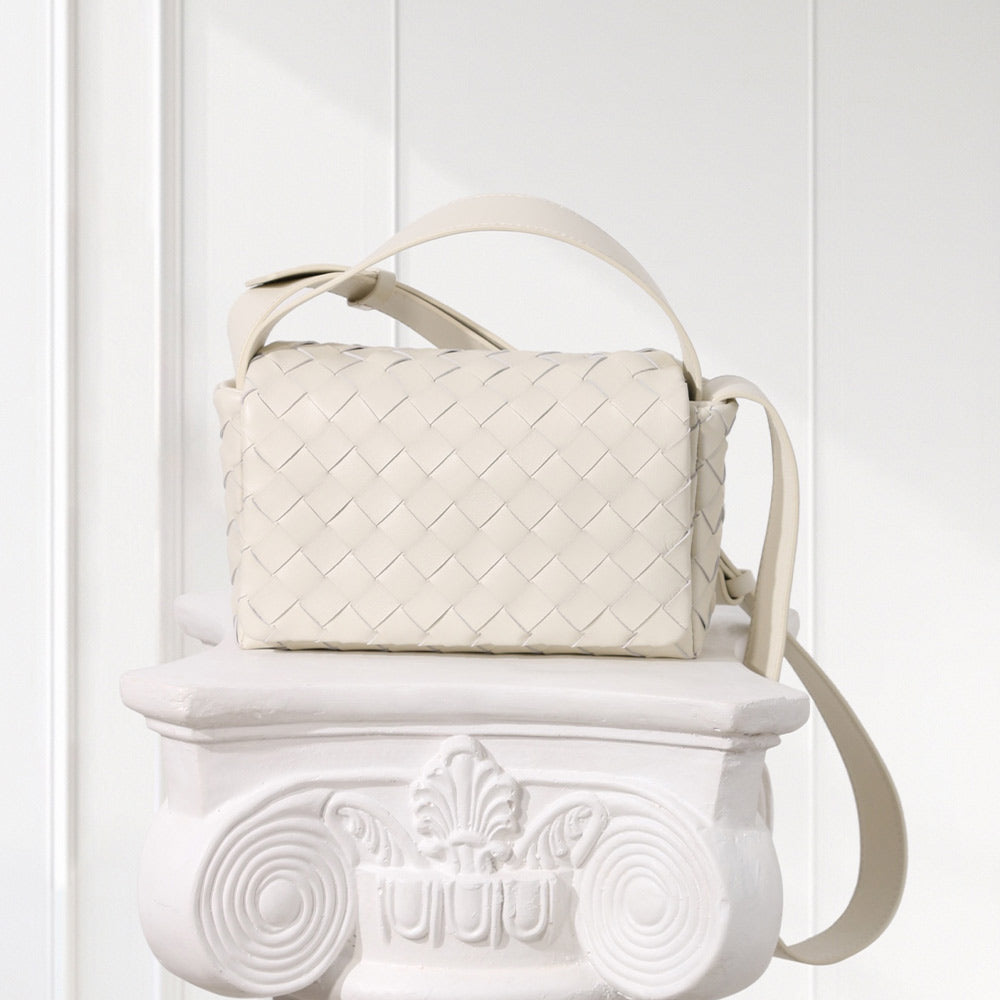 An off-white woven leather shoulder bag with a top handle and adjustable strap, displayed on a white decorative pedestal against a white paneled background.