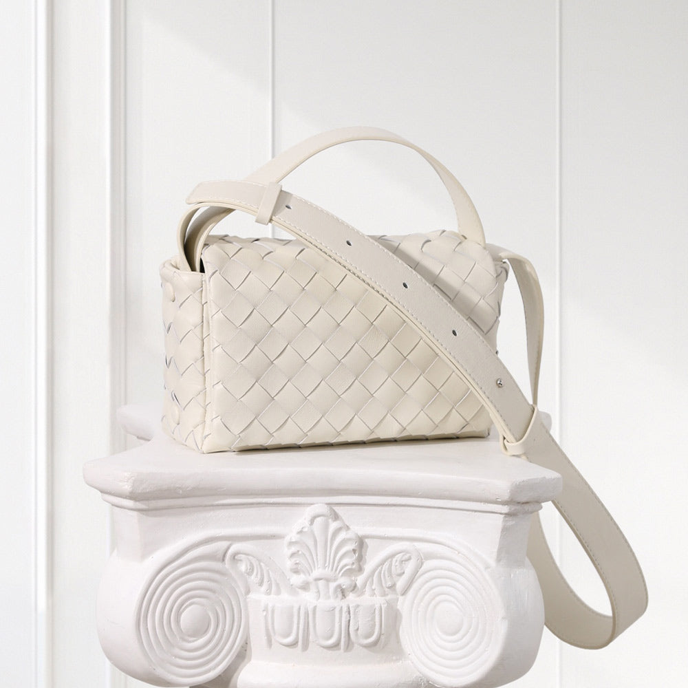 An off-white woven leather handbag with a top handle and shoulder strap, displayed on a white decorative pedestal against a white paneled background.