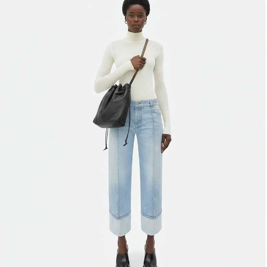 Person wearing a white turtleneck and light blue jeans, holding a black bag on a white background