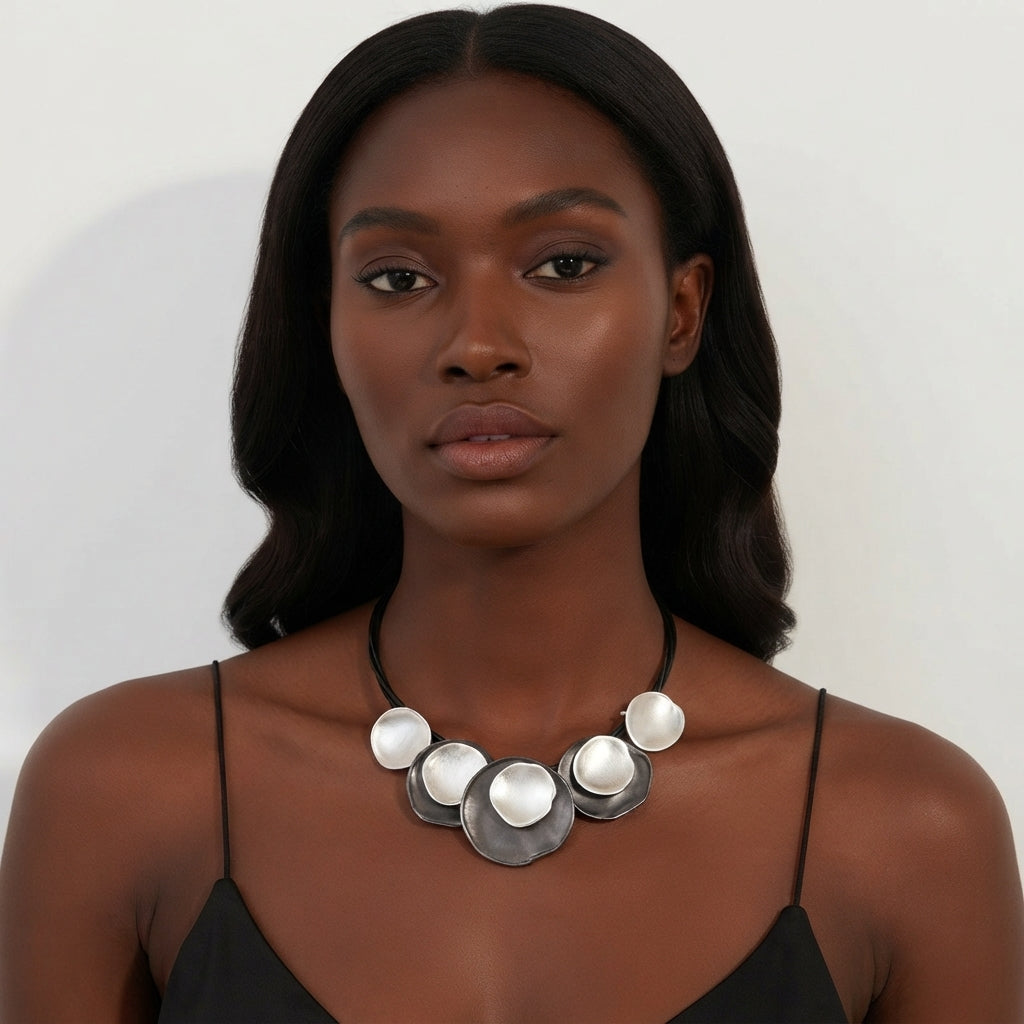A woman with dark hair wearing a black spaghetti-strap top and a chunky silver statement necklace with circular discs, photographed against a plain light background.