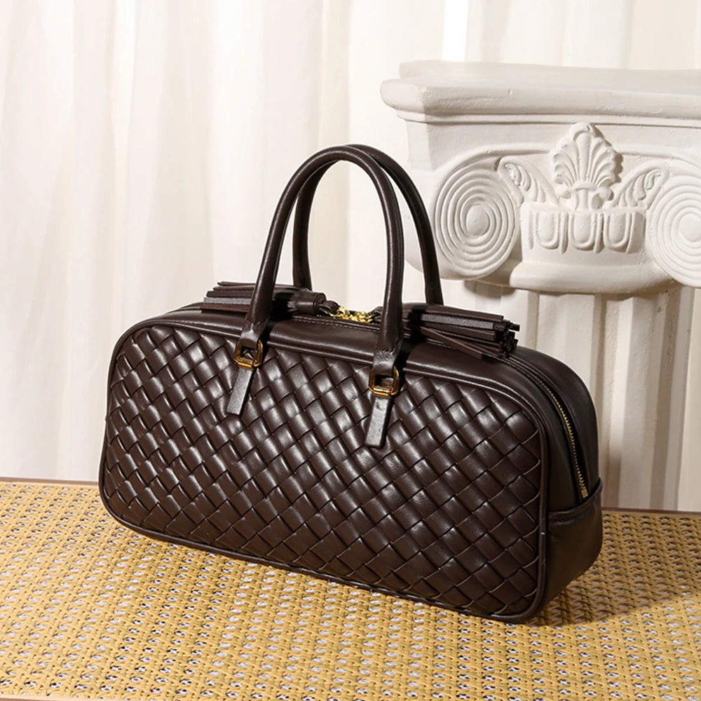 A dark brown quilted leather travel bag with double top handles and gold-tone hardware, displayed on a woven yellow mat in front of a white classical column.